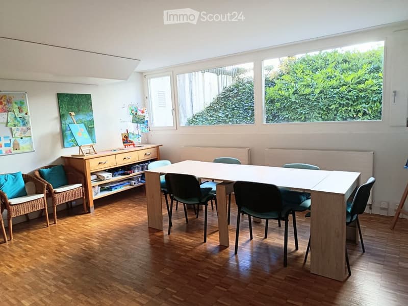 Bright room with wooden flooring, large windows, a long table surrounded by chairs, a wicker sofa with cushions, a wooden cabinet, and several artworks displayed on the walls.