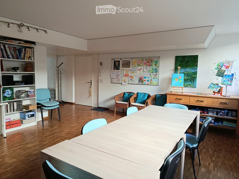 Bright room with wooden flooring, large white table surrounded by light blue chairs, wicker chairs with blue cushions, bookshelf with books and decorative items, easel with painting, and artwork displayed on walls.