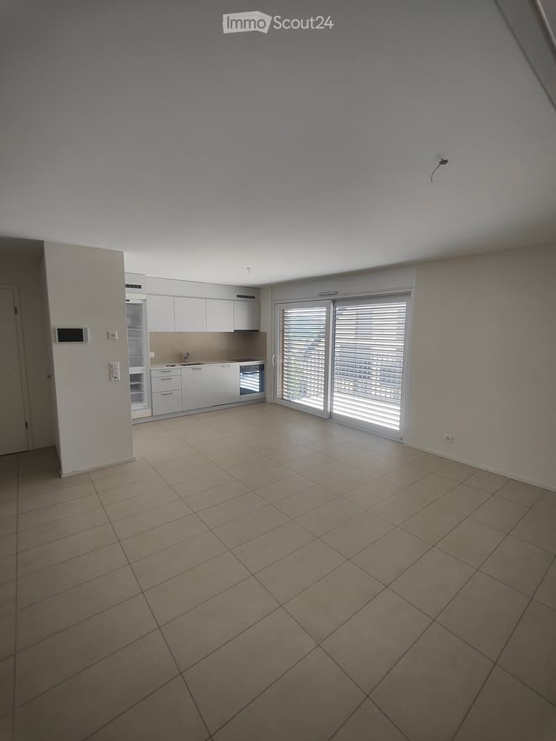 Empty room with tiled floor, white walls, and a small kitchen area with cabinets and appliances visible in the background.
