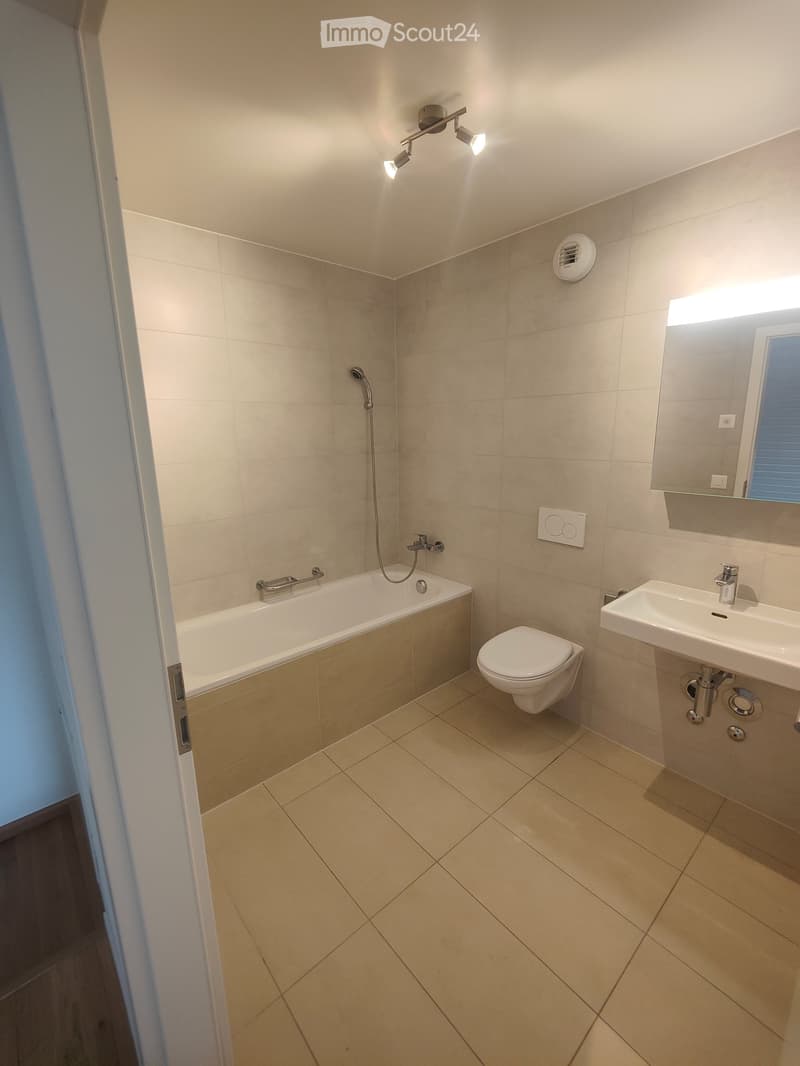 Modern bathroom with beige tiles, a bathtub, toilet, sink, and a mirror with built-in lighting.