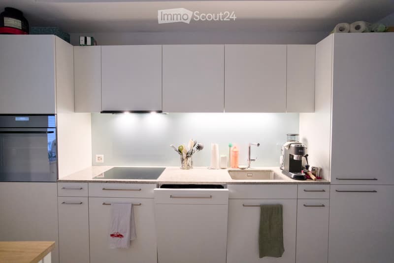 Modern kitchen with white cabinets, stainless steel appliances, and a light-colored countertop. Features include an oven, sink, coffee maker, and a dishwasher.