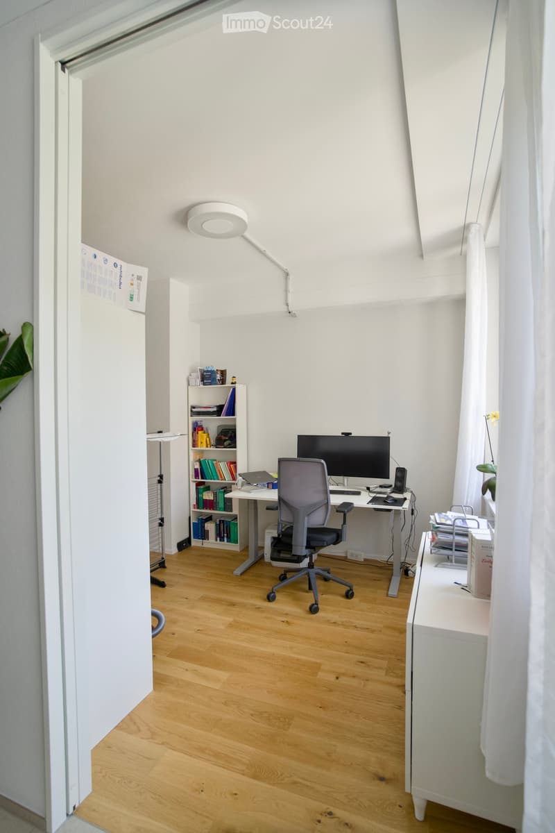 A modern, well-lit office space with a wooden floor, white walls, and large windows. The room features a desk with a laptop, a large screen, and a comfortable office chair. A bookshelf filled with books and decorative items is visible, along with a standi