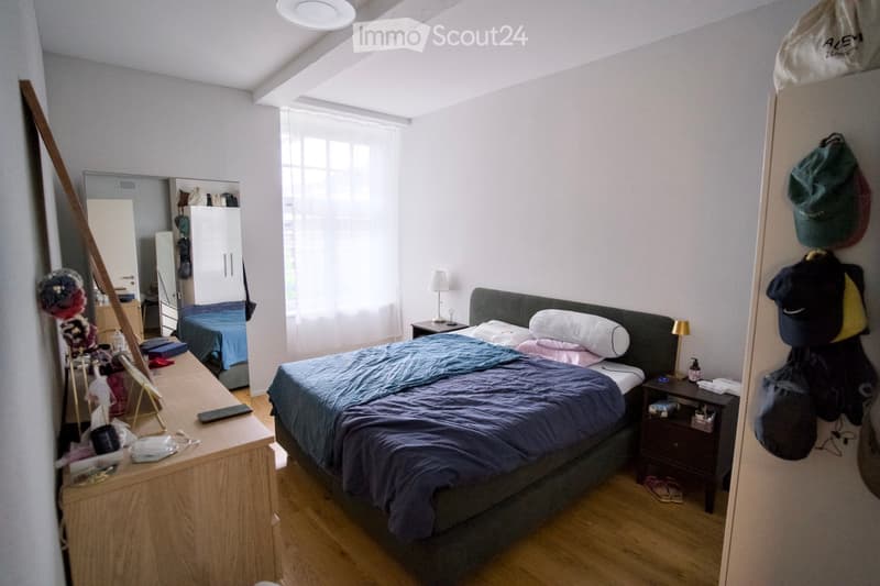 Cozy bedroom with a double bed, nightstands, a mirror, and a wardrobe. The room features wooden flooring and large windows with blinds.