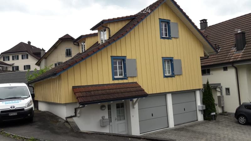 Two-story house with yellow siding, blue-framed windows, and a garage. The house has a sloped roof and is situated in a residential area with neighboring houses.