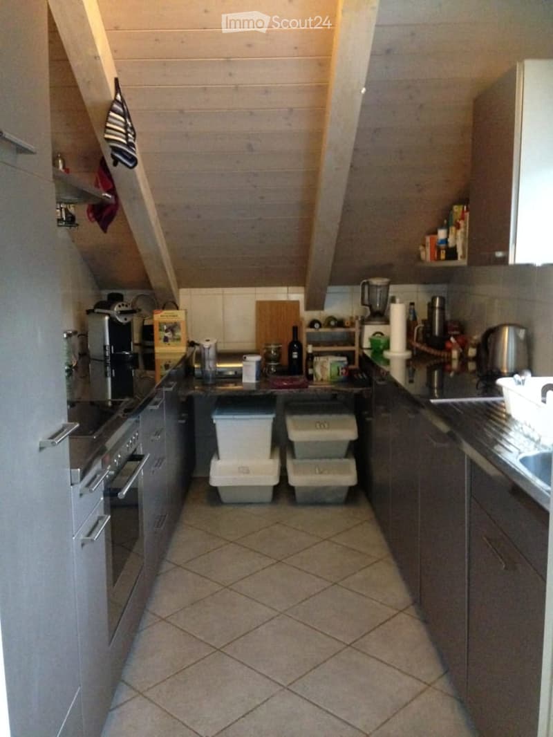 A compact kitchen with grey cabinets, a tiled floor, and various appliances including an oven, refrigerator, and blender. There are also trash bins, a cutting board, and a bottle on the counter.