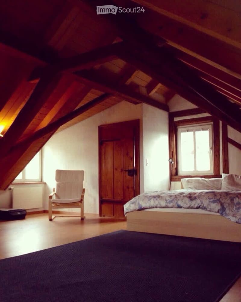 Cozy attic bedroom with wooden beams, slanted ceiling, large windows, bed, chair, and dark blue rug.
