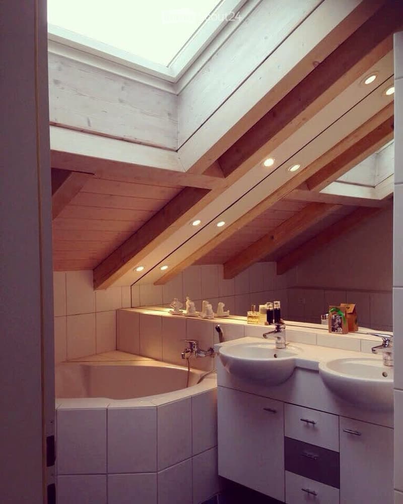 Modern bathroom with skylight, wooden ceiling beams, white tiled walls, double sink vanity, bathtub, and built-in storage cabinets.