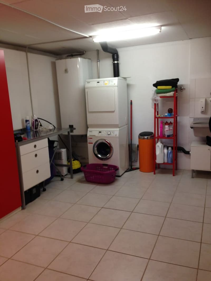 Laundry room with tiled floor, white walls, washing machine, dryer, storage shelf, laundry basket, trash bin, and cabinet.