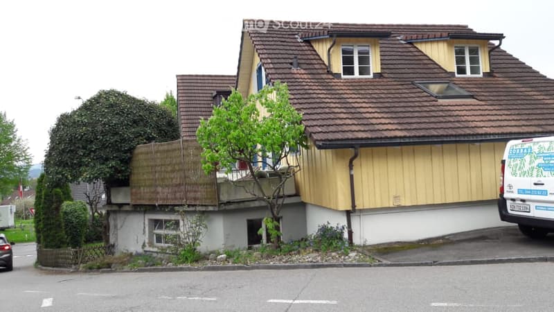 Two-story house with a brown tiled roof, yellow siding, and white trim around windows. The house has a raised ground floor with a small garden area and a parked van in front.