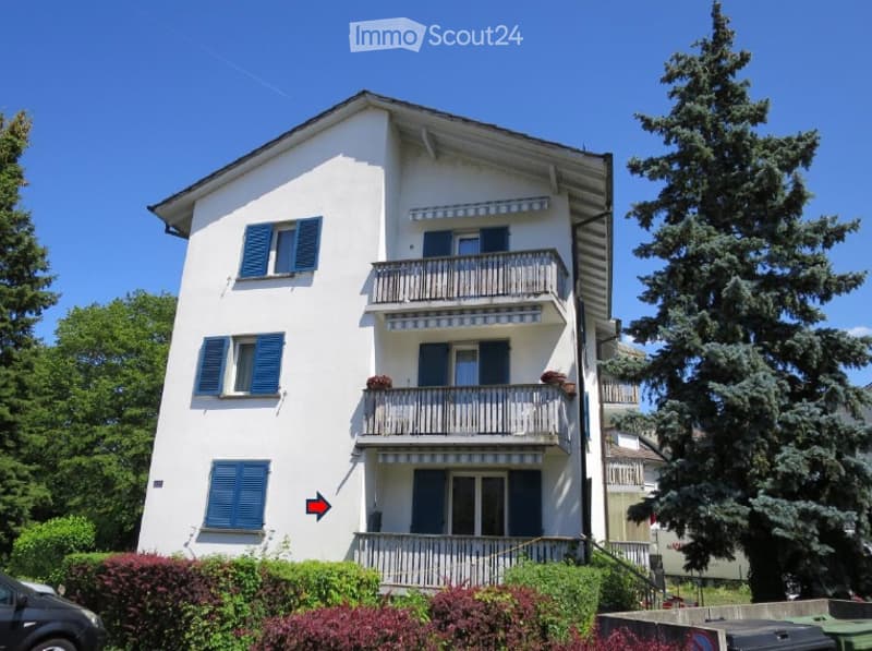 A three-story white building with blue shutters and balconies, surrounded by greenery and trees.