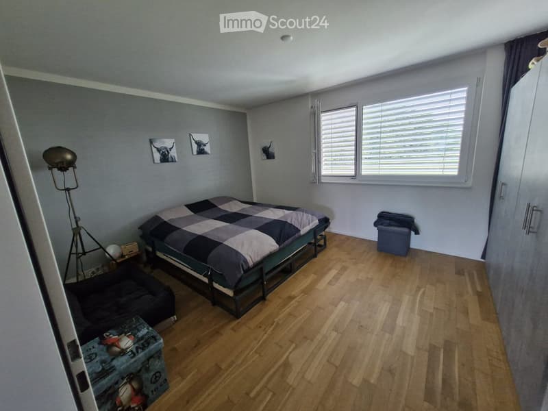 Bedroom with wooden flooring, a bed with a checkered duvet, a nightstand, a floor lamp, and a stool near the window.