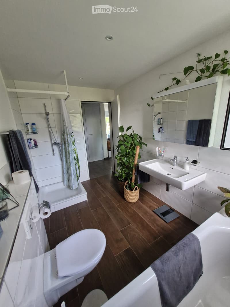 Modern bathroom with wooden flooring, white tiles, a toilet, sink, bathtub, and a large mirror. Includes towels, a plant, and a shower area with a curtain.