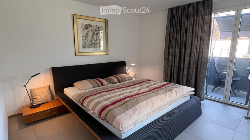 Modern bedroom with a large bed, nightstand, lamp, and a sliding glass door leading to a balcony.