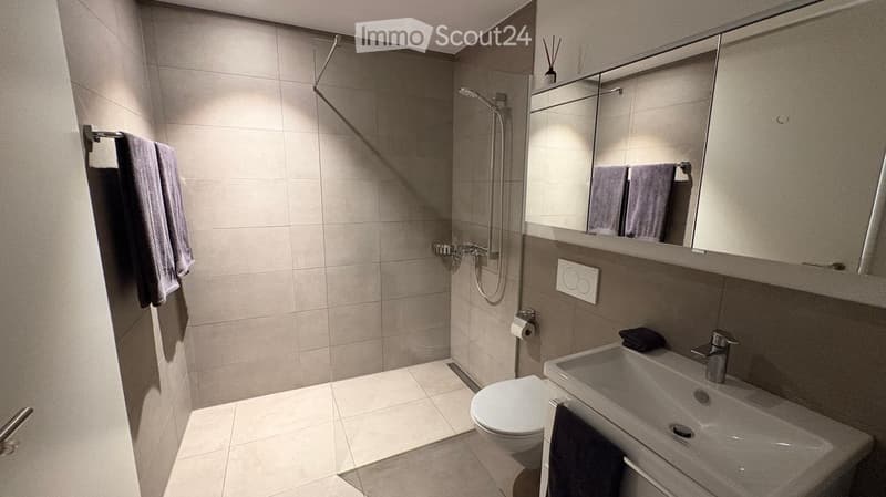 Modern bathroom with a toilet, sink, mirror, and shower. Light gray tiles and a sleek design.