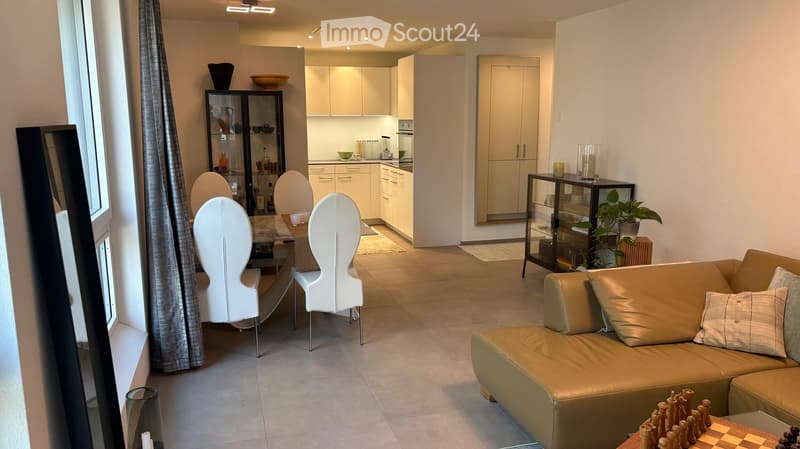 Modern living and dining area with a beige sofa, white chairs, a glass coffee table, and a dining table with four white chairs. The kitchen features white cabinets and a countertop with various appliances.