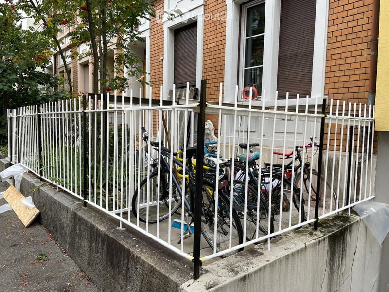 A white metal fence encloses a small outdoor area with several bicycles parked inside. The fence is attached to a concrete platform, and the background shows a multi-story brick building with windows and shutters.