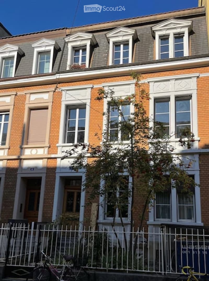A three-story brick building with white decorative trim around the windows and roofline. The facade features multiple windows with white frames and shutters, and a small tree is planted in front of the building.
