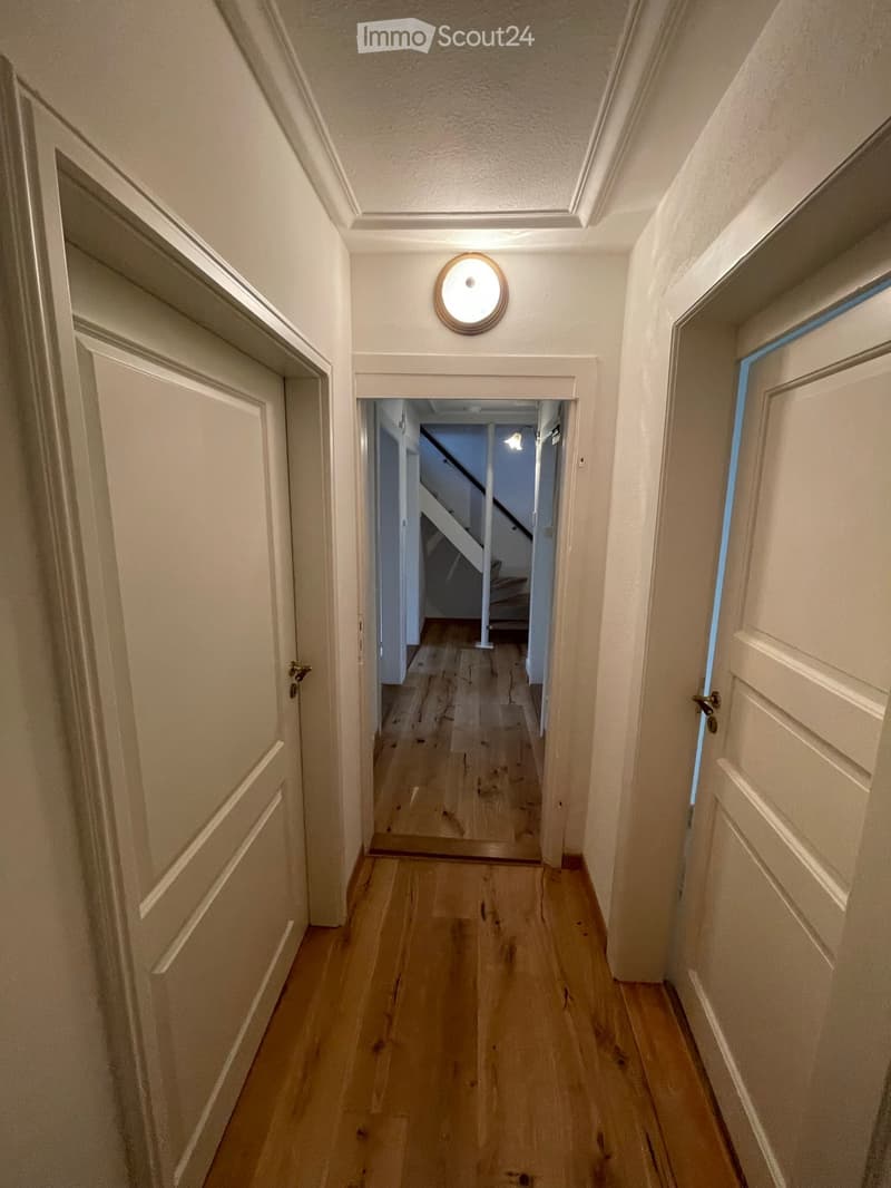 A narrow hallway with wooden flooring, white walls, and several closed doors on both sides. A round wall-mounted light fixture is visible, and a staircase can be seen at the end of the hallway.