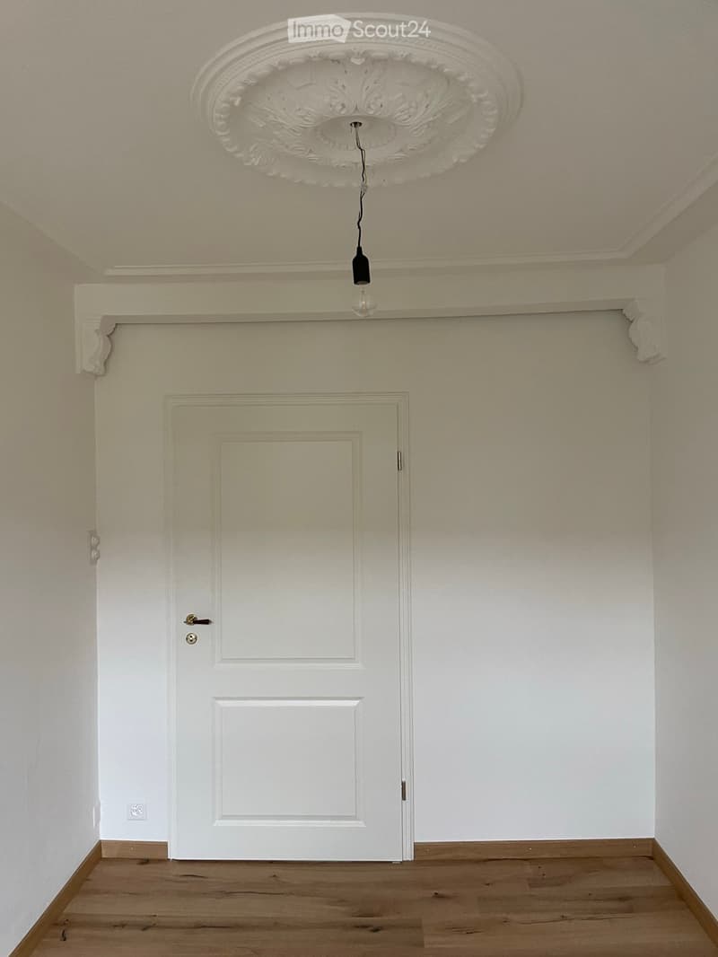 Empty room with light wooden flooring, white walls, a white door, and a decorative ceiling light fixture.