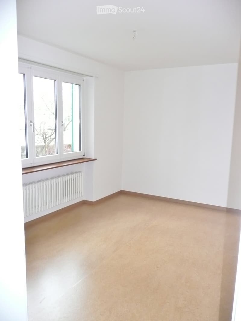 Empty room with white walls, beige tiled floor, large window with white frame, radiator, and wooden shelf.