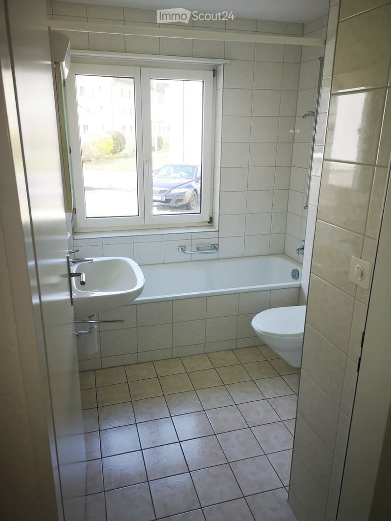 Bathroom with tiled walls and floor, bathtub, toilet, sink, and a window with a view of a parked car.