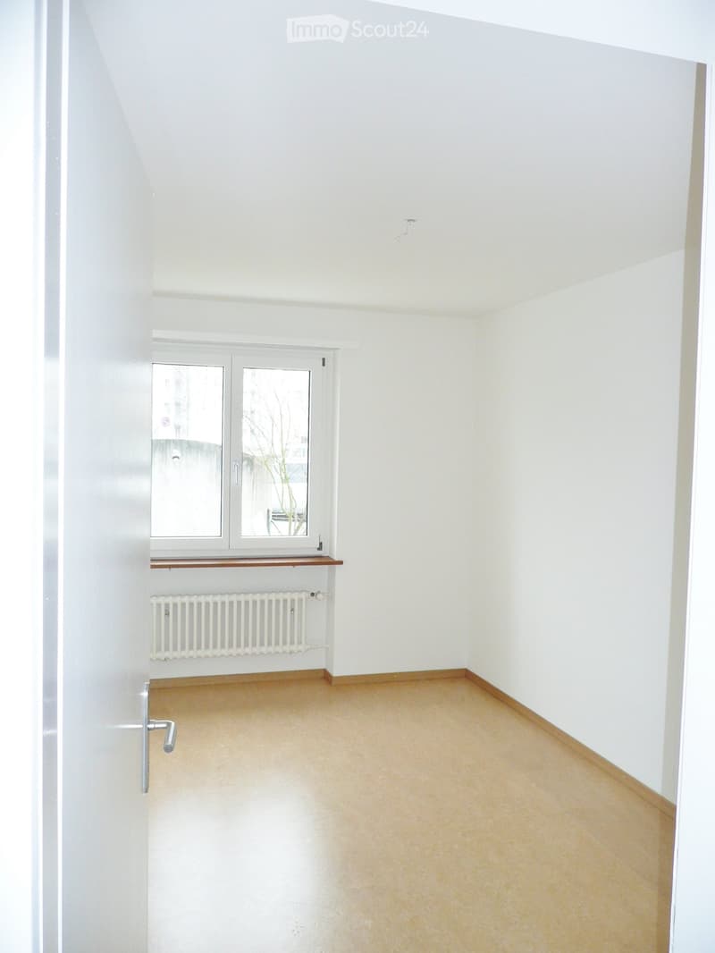 Empty room with white walls, wooden floor, window with double glass panes, radiator, and a small wooden shelf.