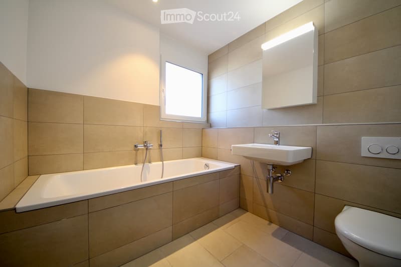 Modern bathroom with beige tiles, a bathtub, a sink, a toilet, and a mirror with built-in lighting.