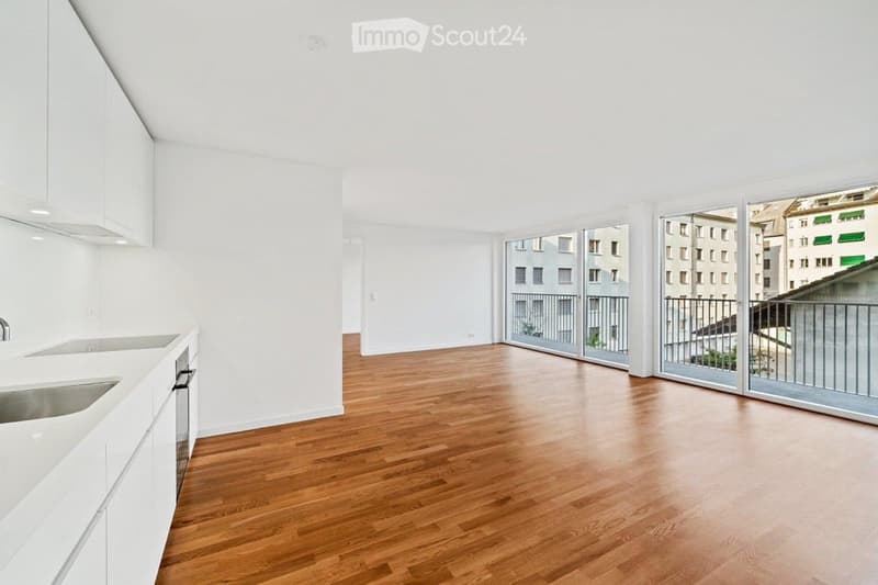 Open-plan unfurnished room with light wooden flooring, white walls, and large sliding glass doors leading to a balcony.