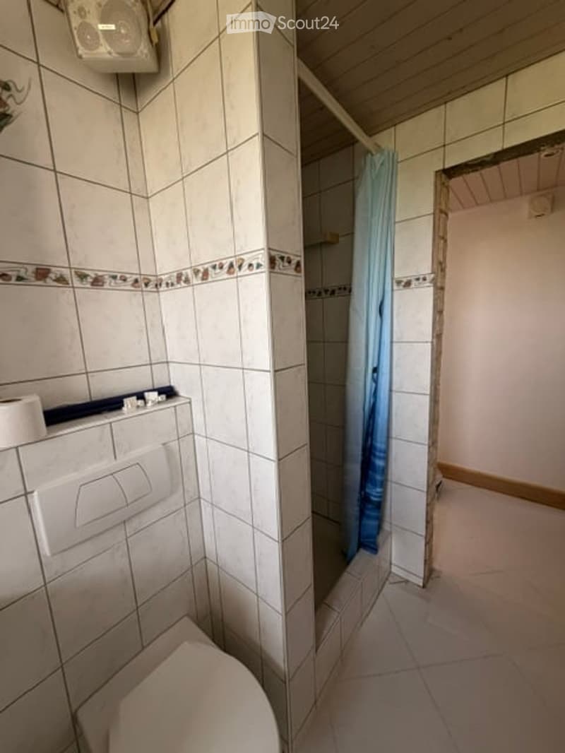 A small bathroom with white tiled walls and floor, a toilet, a shower with a blue curtain, and a toilet paper holder on the wall.