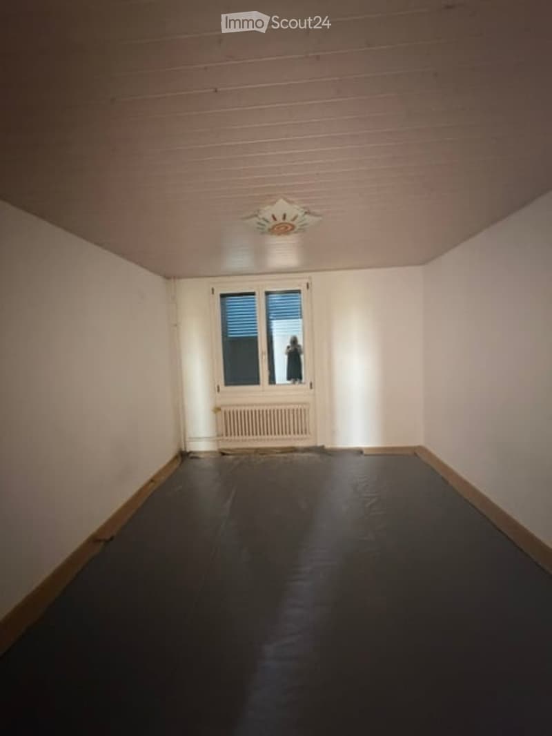 Empty room with white walls, dark flooring, a single window with blinds, a radiator, and a ceiling light fixture.
