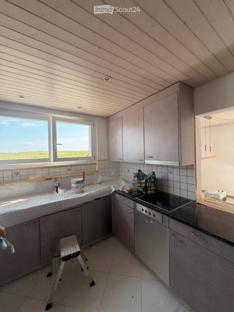 Modern kitchen with light grey cabinets, white countertops, and stainless steel appliances. Features a sink, dishwasher, stove, and a small stool near the counter.