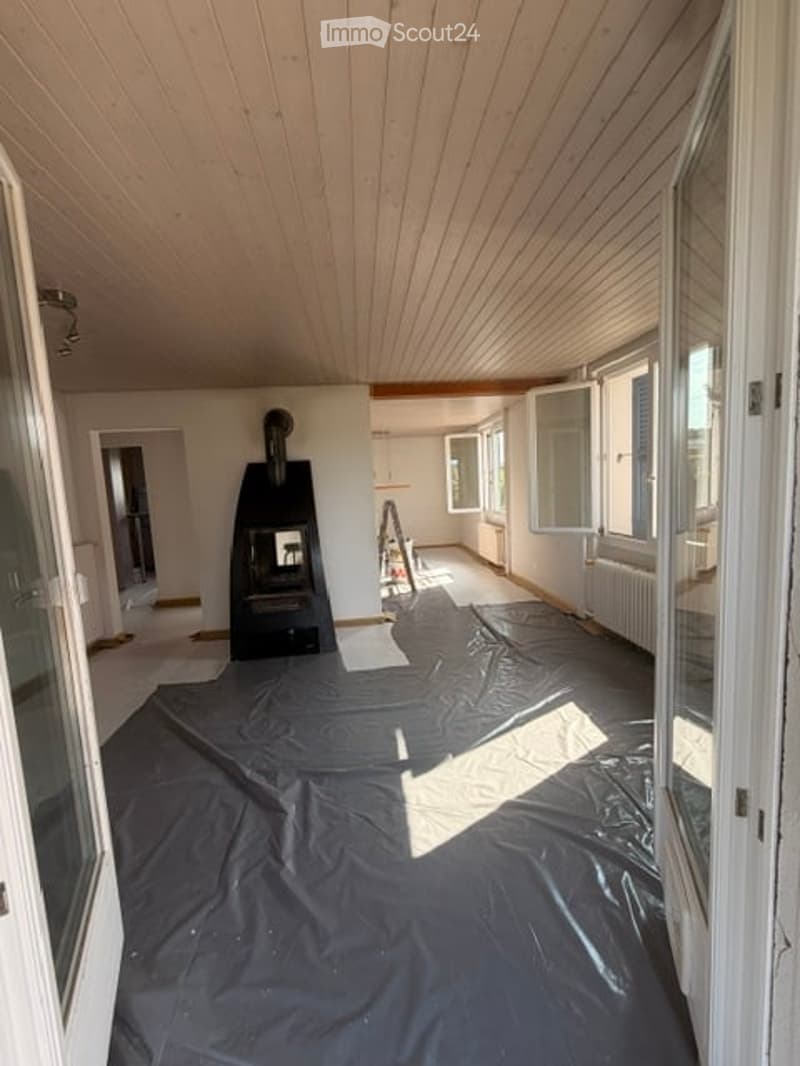 Empty room with wooden ceiling, large windows, and a fireplace. Floor covered with protective plastic sheeting.