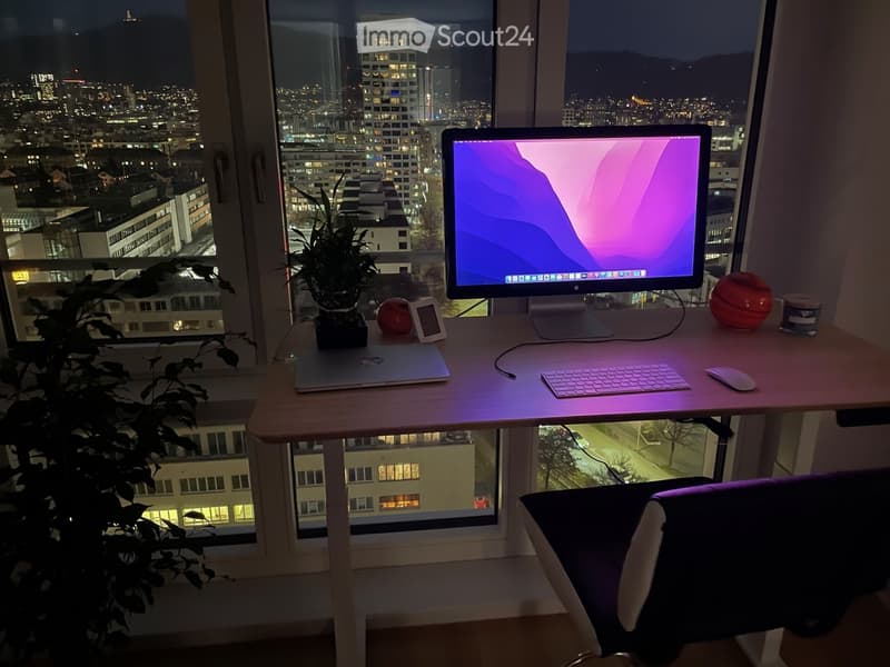 A modern workspace with a desk, laptop, monitor, keyboard, mouse, chair, and a potted plant, overlooking a cityscape at night.