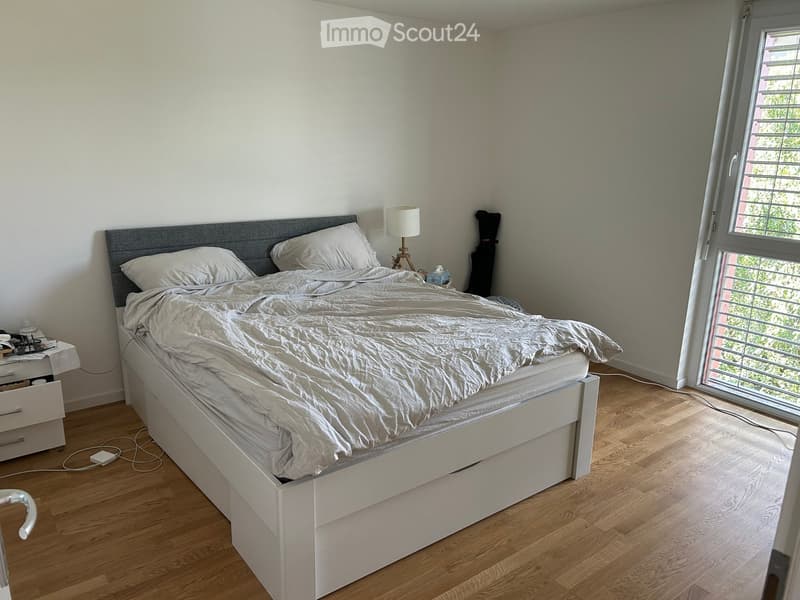A modern bedroom with a white bed, grey headboard, and a nightstand with a lamp. The room features light wooden flooring and a large window with blinds.
