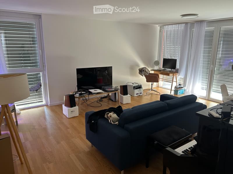 Living room with wooden flooring, large windows, a blue sofa, a piano, a table with a screen, and a chair.