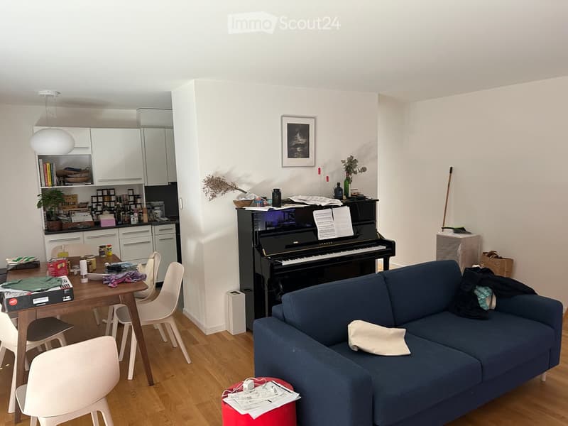 A modern, open-plan living and dining area with a blue sofa, wooden dining table, white chairs, a black piano, and a small kitchen area with white cabinets and a countertop.