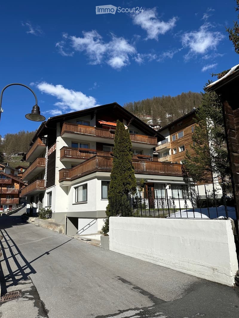 Multi-story building with balconies, wooden railings, and a stone facade, surrounded by trees and a clear blue sky.