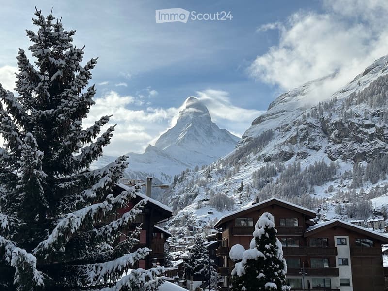 Snow-covered village with traditional wooden houses and snow-laden trees, set against a backdrop of snow-capped mountains.
