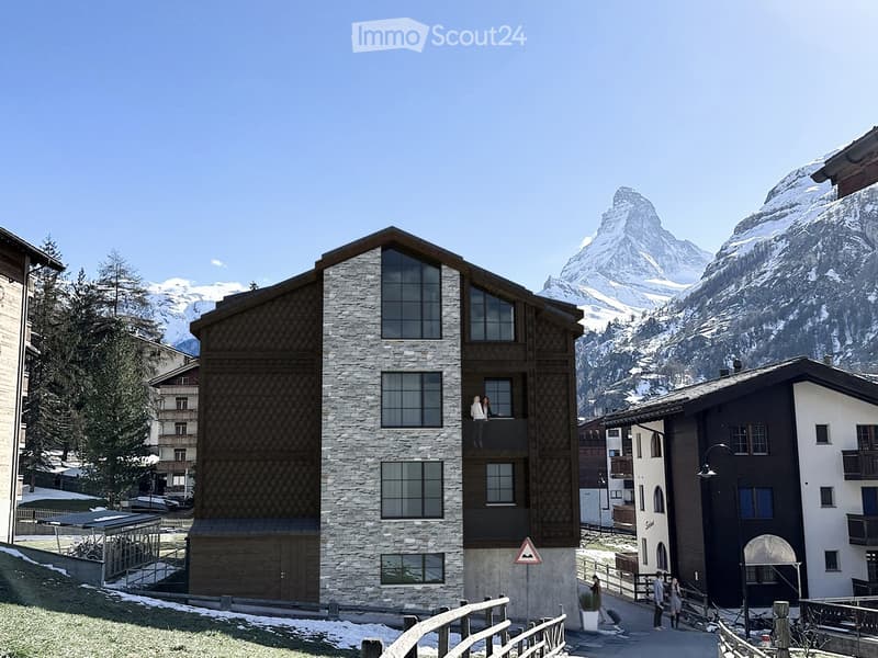 A modern stone and wood building with large windows and balconies, situated in a picturesque alpine village with snow-capped mountains in the background.