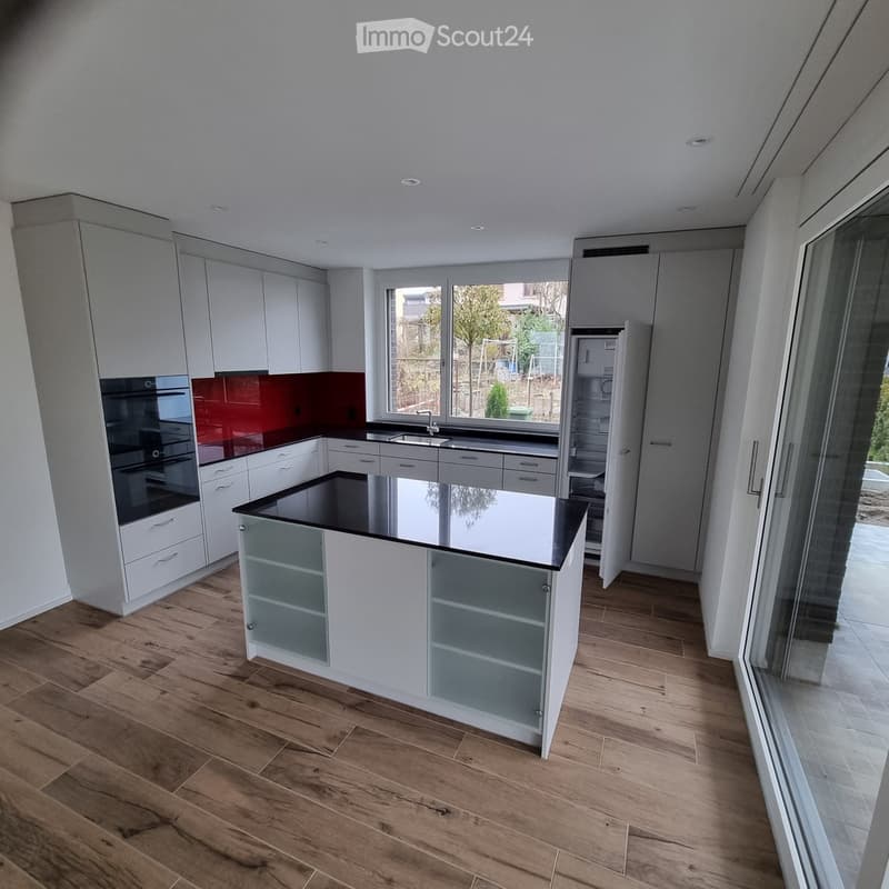 Modern kitchen with white cabinetry, a central island with glass-fronted storage, a black countertop, and a red backsplash. Features include a sink, faucet, oven, and refrigerator, with large windows offering a view of the garden.