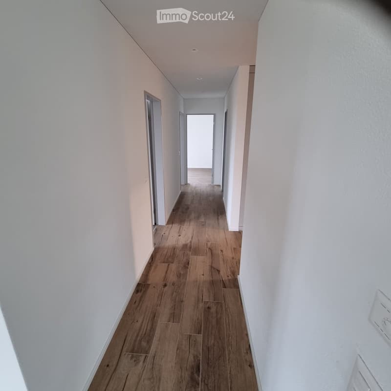 A clean, well-lit hallway with wooden flooring and white walls, featuring several doors and a bright opening at the end.