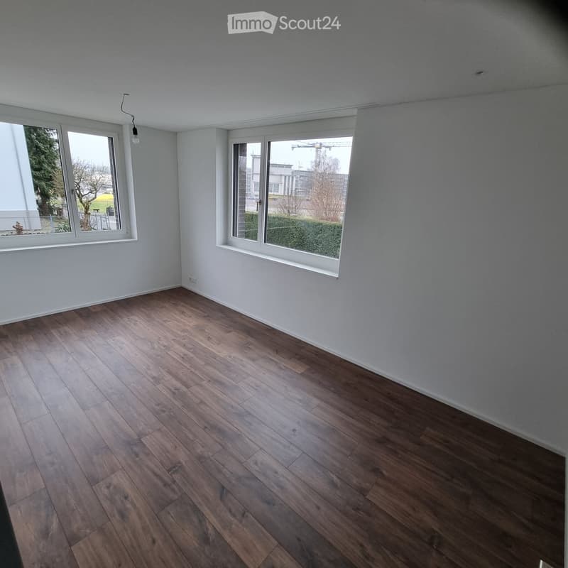 Empty room with wooden flooring, white walls, and large windows offering a view of the outside area.
