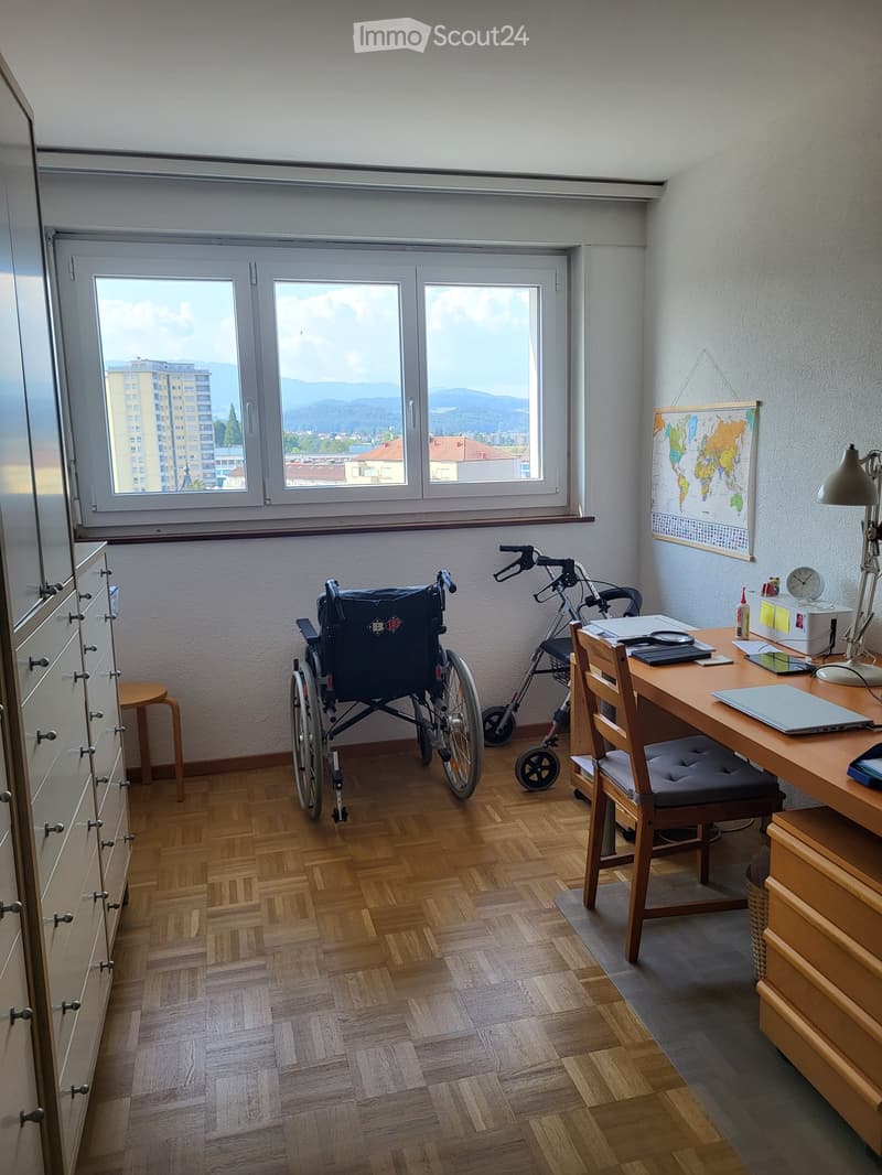 A furnished room with a wheelchair, walking aid, desk, chair, laptop, lamp, cabinet, stool, map, and clock. The room has wooden flooring and large windows with a scenic view.