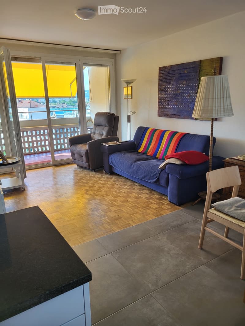 Living room with a blue sofa, a brown armchair, a wooden chair, a floor lamp, and an art piece on the wall. The room features a balcony with a view and a mix of wooden and tiled flooring.