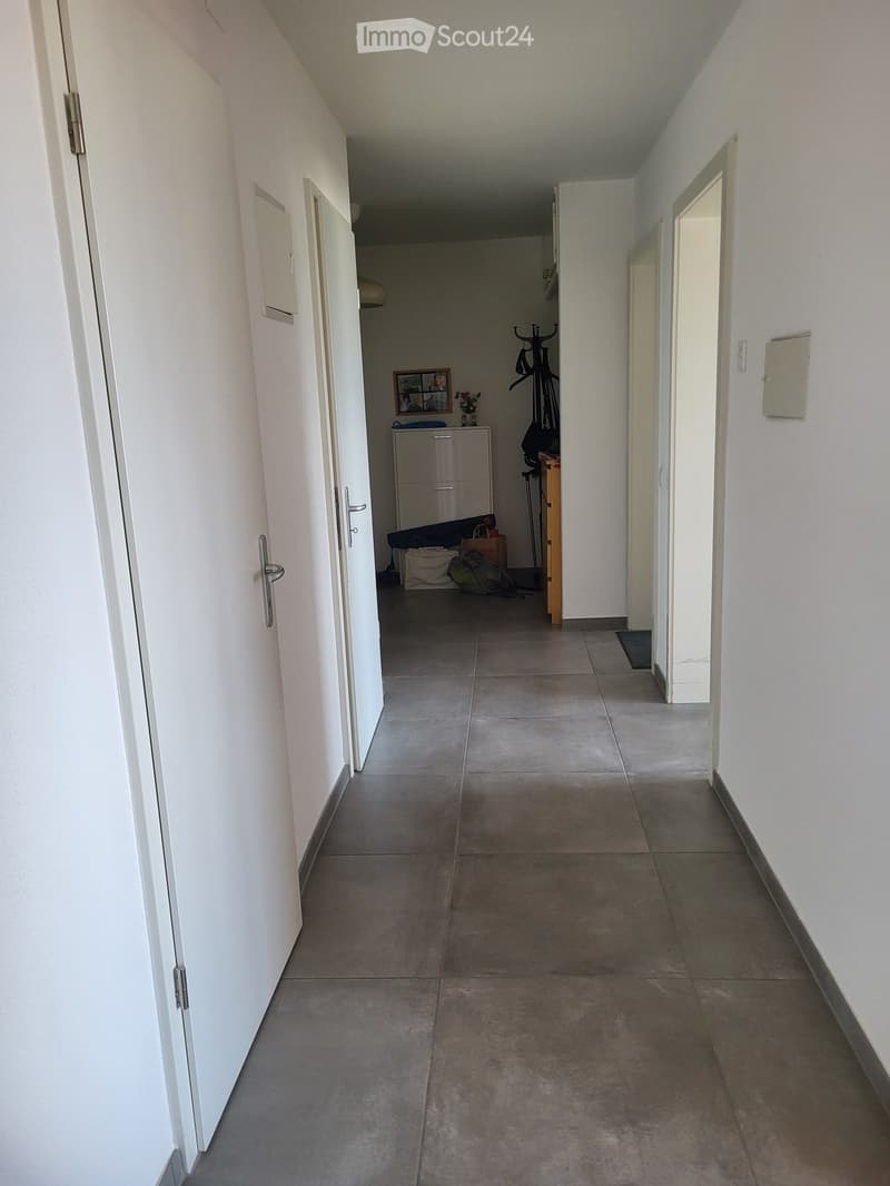 A long, narrow hallway with tiled flooring and white walls. Doors are visible on both sides, and there is a small area with a cabinet, a bicycle, and some bags at the end of the hallway.