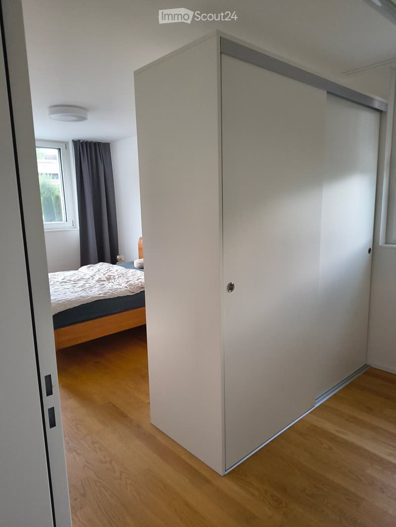 A modern bedroom with wooden flooring, a bed with a white duvet, a nightstand with a lamp, and a large window with dark curtains.