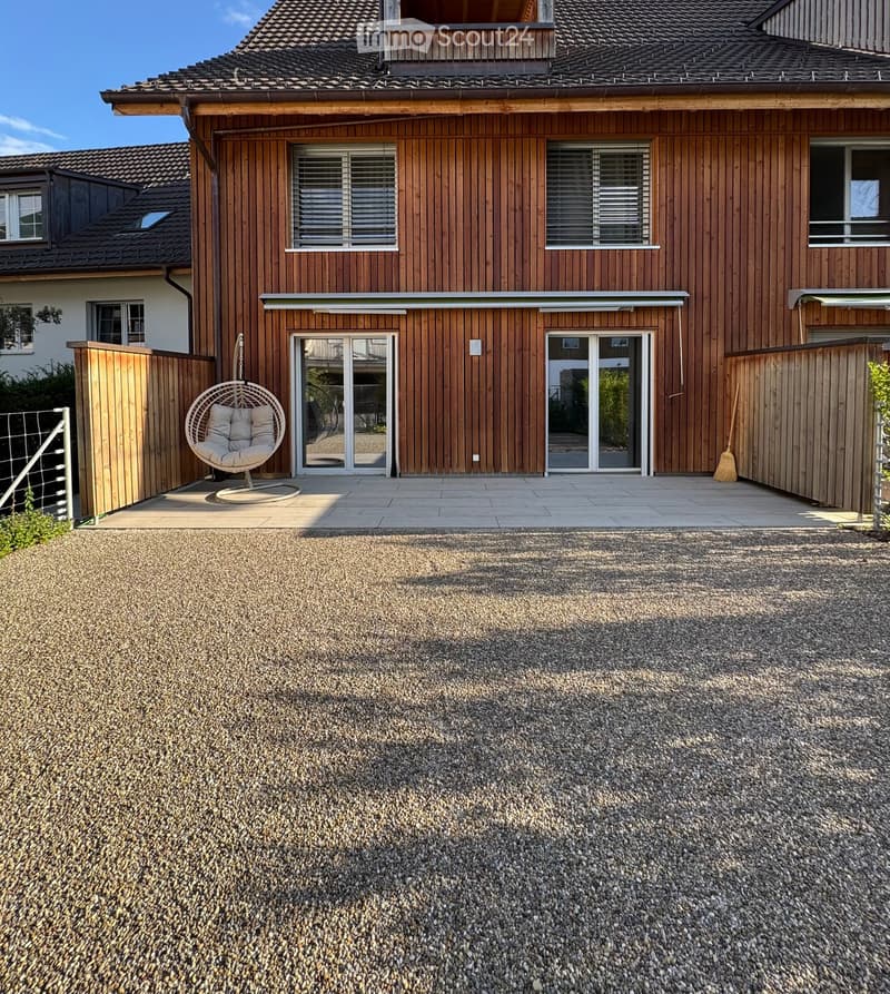 Modern wooden house with large windows and sliding glass doors, featuring a gravel driveway and a wicker hanging chair on the patio.