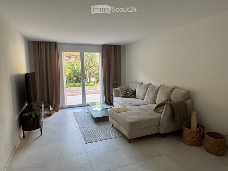 Modern living room with a grey sectional sofa, glass coffee table, area rug, and wicker baskets. Large sliding glass doors lead to a patio.