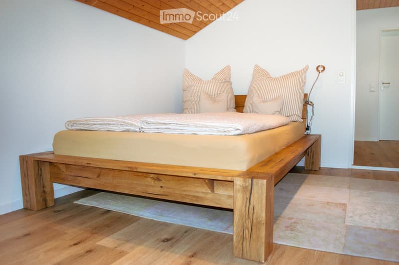 Cozy bedroom with a wooden bed, striped pillows, and a warm-toned blanket. The room features wooden flooring and a slanted wooden ceiling.