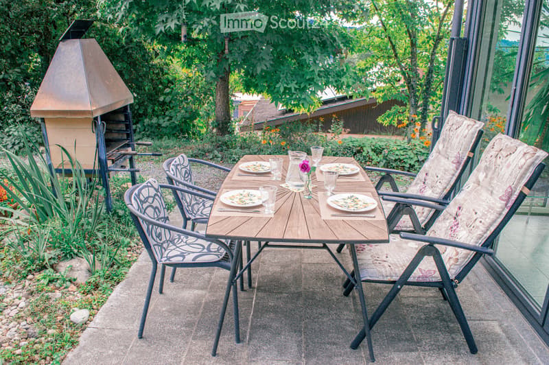 Outdoor patio area with a wooden dining table, four chairs, plates, glasses, and a vase with a flower. A barbecue grill is visible to the left, surrounded by greenery and plants.